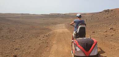 Frau mit Fahrrad, Kindersitz und Anhänger fährt durch eine wüstenartige Landschaft