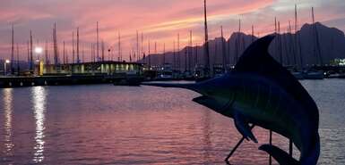 Schwertfischstature im Meer, dahinter Segelboote und  rosafarbene Wolken